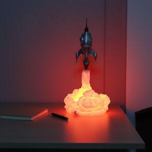 Peut inclure: Lampe fusée rétro argentée avec une base nuageuse lumineuse. La lampe émet une lumière orange chaude. Un stylo et un carnet sont sur la table. La lampe mesure environ 30 cm de haut.