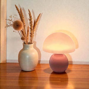 May include: A pink mushroom-shaped table lamp with a rounded base and a semi-circular top. The lamp is illuminated, casting a warm glow. Beside it, a cream-colored vase holds dried flowers and wheat stalks, creating a cozy, decorative scene.