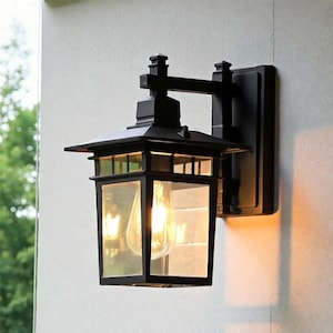 Puede incluir: Lámpara de pared exterior negra con una pantalla rectangular con paneles de vidrio. La linterna presenta un diseño clásico con un techo puntiagudo y una bombilla visible. El accesorio está montado en una placa trasera cuadrada.