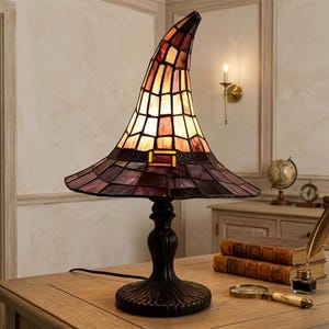 May include: A stained glass table lamp shaped like a pointed witch hat. The lamp features a dark purple brim and a lighter, warm-toned crown with a rectangular buckle detail. It stands on a dark, ornate, pedestal-style base with a textured finish.