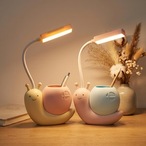 Puede incluir: Dos lámparas de escritorio caprichosas con forma de caracol con portalápices. Una es amarilla y rosa, la otra rosa y azul. Cada una tiene un cuello blanco flexible y una luz rectangular. Un bolígrafo y un lápiz están en los soportes.
