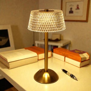 May include: A small, gold-coloured table lamp with a diamond-patterned lampshade. The lamp has a round base and a slender stem. The lamp is on a white surface with books and a pen nearby. The lamp is designed for indoor use.