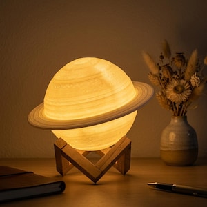 May include: A Saturn-shaped lamp with a detailed ring system, emitting a warm glow. The lamp sits on a wooden stand. The lamp is a decorative item, perfect for a bedroom or living room. The lamp is a decorative item, perfect for a bedroom or living room.