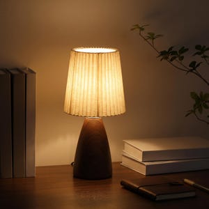 May include: A table lamp with a pleated lampshade and a wooden base. The lampshade is beige and the base is a dark brown, conical shape. The lamp is on a wooden surface with books and a notebook. The lamp is lit, casting a warm glow.