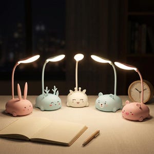 May include: A collection of five whimsical desk lamps in the shape of animals, including a bunny, deer, giraffe, bear, and pig. Each lamp has a flexible neck and a bright, oval-shaped light. The lamps are in pastel colors and sit on a desk.