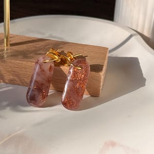 Könnte beinhalten: Ein Paar Ohrhänger mit goldfarbenen Haken. Die Ohrringe sind rotbraun mit Goldflitter. Die oval geformten Ohrringe liegen auf einem Holzblock.