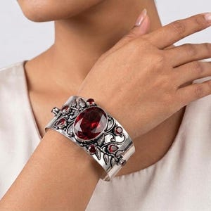 May include: A wide silver cuff bracelet featuring an ornate metalwork design of leaves and vines. The centre is set with a large, oval-shaped deep red gemstone, surrounded by several smaller, round and teardrop-shaped red stones embedded in the metal.