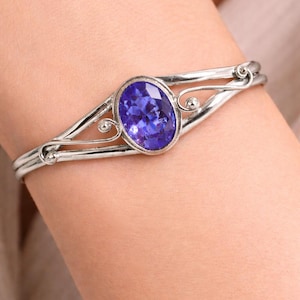 Può includere: Un bracciale color argento con una gemma viola sfaccettata di forma ovale al centro. La fascia in metallo presenta un design diviso con delicati dettagli in filo metallico a spirale su entrambi i lati della pietra, creando una struttura aperta.