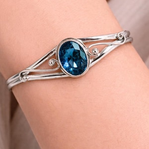 May include: A silver-toned cuff bracelet featuring a central oval-shaped faceted blue gemstone. The metal band splits into two delicate, curved arms on each side of the stone, accented with small spherical metal beads at the terminal points.