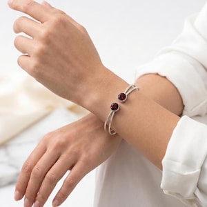 May include: A silver-toned open cuff bracelet featuring two round, deep red stones set at each end. The bracelet is constructed from two parallel thin metal bands that meet at the stone settings, creating a sleek and minimalist design.