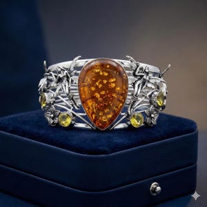 May include: A silver cuff bracelet with a large amber stone, flecked with gold. The bracelet features fairy figures, flowers, and yellow gemstones. The jewellery is displayed on a dark blue velvet surface, creating a striking contrast.