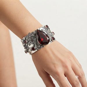 May include: A wide silver cuff bracelet featuring a large, central, pear-shaped dark red gemstone. The metal surface is decorated with intricate floral and scrollwork engravings, accented by several smaller, round, dark red gemstones.