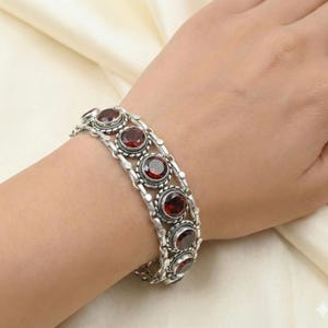 Può includere: Un bracciale in argento caratterizzato da una serie di pietre preziose granato rotonde e rosso scuro, incastonate in castoni di metallo decorati e testurizzati. Il bracciale è composto da più segmenti d'argento collegati che formano una fascia.