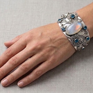 May include: A wide silver cuff bracelet featuring an oval iridescent moonstone centre stone surrounded by intricate floral metalwork and small round blue gemstones set throughout the ornate, polished silver band design.