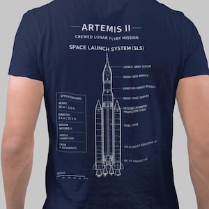 Puede incluir: Una camiseta azul marino con un diagrama técnico blanco del cohete Space Launch System Artemis II en la parte posterior. El gráfico incluye etiquetas para los componentes del cohete, especificaciones de la misión y una escala en metros.