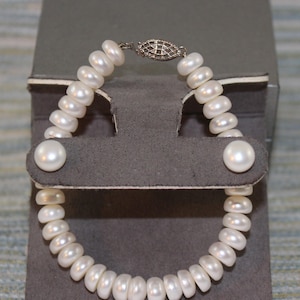 Può includere: Un braccialetto composto da numerose perle d'acqua dolce bianche a forma di disco infilate insieme. Il pezzo presenta una chiusura decorativa in metallo color argento di forma ovale con un intricato motivo a reticolo che unisce le estremità.