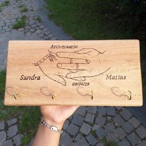 Puede incluir: Un soporte de pared rectangular de madera clara para llaves con una ilustración grabada a láser de dos manos entrelazadas. El texto dice Aniversario, Sandra, Matias y 09/04/25. Incluye cuatro ganchos de metal en tono dorado.
