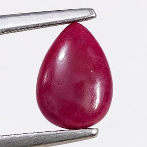 May include: A pear-shaped, deep red gemstone with a smooth, polished surface. The stone is held between the metal tips of a pair of tweezers, showcasing its vibrant color and teardrop silhouette against a plain, light-colored background.