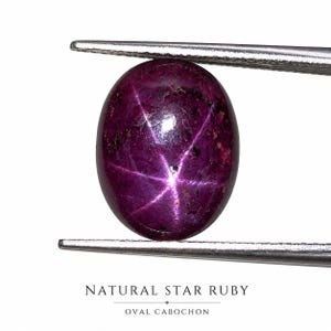 Könnte beinhalten: Ovalförmiger natürlicher Sternrubin-Edelstein, gehalten von einer Pinzette. Der tiefmagentafarbene Stein zeigt ein sechsstrahliges Sternmuster. Der Text unten lautet "NATURAL STAR RUBY" und "OVAL CABOCHON".