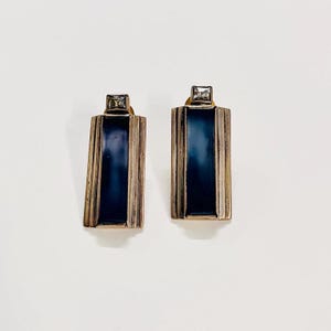 Pode incluir: Um par de brincos em estilo Art Déco. Cada brinco apresenta um centro retangular de esmalte azul escuro emoldurado por metal dourado. Um pequeno cristal transparente quadrado cobre cada brinco. Em um fundo branco.