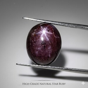 Könnte beinhalten: Ein ovaler, hochwertiger natürlicher Sternrubin-Edelstein, der von einer Pinzette gehalten wird. Der Rubin hat eine tief burgunderrote Farbe mit einem sechsstrahligen Sternmuster. Der Text "HIGH-GRADE NATURAL STAR RUBY" befindet sich unten.