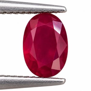 Könnte beinhalten: Ein leuchtend tiefroter Edelstein im Ovalschliff, der sicher von einer silberfarbenen Metallpinzette gehalten wird. Der Rubin besitzt eine facettierte Oberfläche, die das Licht reflektiert und seine satte karmesinrote Farbe sowie die klare, symmetrische ovale Form betont.