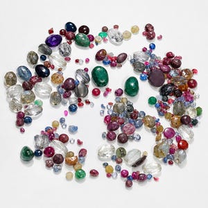 Può includere: Una collezione di pietre preziose sfuse multicolori di varie forme e dimensioni, tra cui pietre trasparenti, verdi, blu, rosse, viola e gialle. L'assortimento presenta sia tagli sfaccettati che cabochon lisci e arrotondati sparsi.