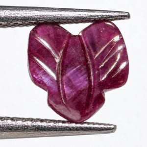 Puede incluir: Una piedra preciosa de color magenta intenso tallada en forma de hoja o pétalo estilizado con surcos verticales distintivos. La piedra translúcida muestra inclusiones naturales y está sujeta por las puntas metálicas de unas pinzas.