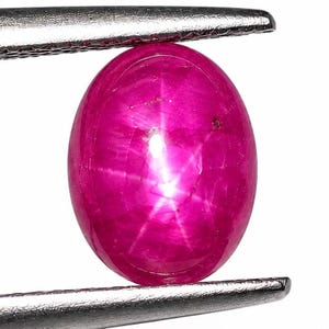 Könnte beinhalten: Ein leuchtender, ovaler Sternrubin-Cabochon in einem tiefen Magentarosa. Der Edelstein zeigt einen ausgeprägten sechsstrahligen Asterismus-Effekt, der durch ein helles, weißes Sternmuster in der Mitte der glatten, polierten und gewölbten Oberfläche.
