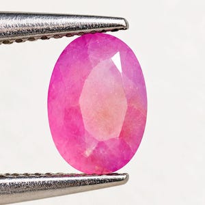May include: An oval-cut, vibrant pink gemstone held by silver-toned metal tweezers. The stone features a faceted surface that reflects light, showcasing a translucent, saturated pink hue throughout its polished, symmetrical oval shape.