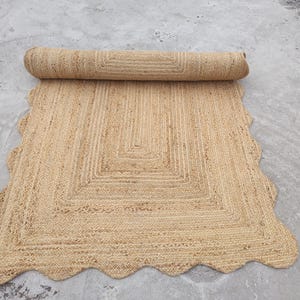 May include: A rectangular area rug crafted from natural tan woven fibers, featuring a rolled top section and a decorative scalloped edge border. The rug displays a concentric rectangular pattern created by the braided texture of the material.