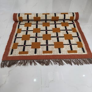 May include: A rectangular area rug featuring a grid pattern of black lines and solid mustard-brown squares on an off-white background. The rug has a wide terracotta-coloured border and a fringe of brown tassels along the bottom edge.