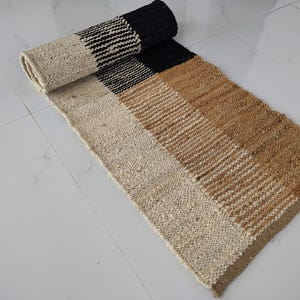 May include: A rectangular woven runner rug featuring a colour-blocked design with sections of cream, black, and tan. The rug has a textured, chunky weave and is partially rolled at one end, displaying a consistent pattern of horizontal stripes.