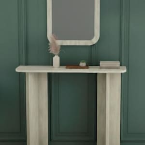 May include: A modern, light-colored console table with a matching mirror. The table has a rectangular top and two vertical supports. A small vase with dried flowers, a tray with a plant, and books are on the table. The mirror is rectangular with rounded corners.