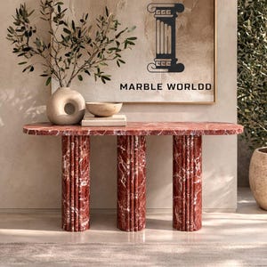 May include: A red marble console table with three fluted cylindrical legs. The tabletop is oval-shaped. The text "MARBLE WORLDD" is visible on the wall behind the table. A vase with greenery and a bowl are on the table.