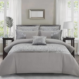 May include: A grey bedspread set featuring a damask pattern and quilted texture. The set includes a comforter, pillows, and shams in various shades of grey. A decorative pillow is placed in the centre of the bed.