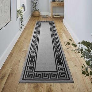 May include: A long, rectangular runner rug featuring a solid light grey centre panel framed by a bold, black Greek key geometric border pattern. The rug is positioned lengthwise on a light-toned wooden floor, extending towards the back of the room.