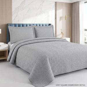 May include: A grey bedspread and two matching pillow shams featuring a textured square embroidery pattern. The bedding set has a uniform light grey colour and a quilted appearance with a repeating geometric square design across the fabric surface.