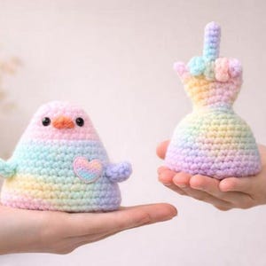 Op de afbeelding: Twee handgemaakte gehaakte figuren: een vogelachtig wezen met een hart en een hand die de middelvinger opsteekt. Beide zijn in pastel regenboogkleuren. De vogel heeft een roze kop, een oranje snavel en een hartvormige patch. De hand heeft de vorm van een jurk.