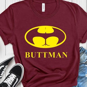 May include: A maroon short-sleeve t-shirt featuring a yellow graphic of a stylized bat-like emblem shaped like buttocks, with the text "BUTTMAN" printed in yellow capital letters underneath the graphic on the front of the garment.