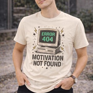 May include: A cream-colored short-sleeve t-shirt featuring a graphic of a vintage computer monitor displaying the green text ERROR 404, with the black text MOTIVATION NOT FOUND printed below the computer, surrounded by small star and lightning bolt icons.