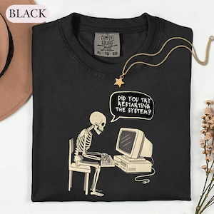 May include: A black t-shirt featuring a graphic of a skeleton sitting on a chair at a computer. The skeleton is typing on a keyboard and a speech bubble above it reads, "DID YOU TRY RESTARTING THE SYSTEM?" The shirt has a Comfort Colors brand label.