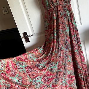 May include: A floor-length halter dress featuring a vibrant paisley pattern in shades of pink, teal, and orange. The garment has a deep V-neckline, a gathered waistline, and a full, flowing skirt that drapes to the floor with a wide hemline.