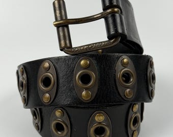 Puede incluir: Un cinturón de cuero negro con una serie de placas metálicas ovaladas en tono latón, cada una con un ojal central y dos remaches pequeños. El cinturón incluye una hebilla rectangular en tono latón con el texto Dolce & Gabbana grabado.
