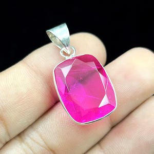 May include: A rectangular pendant featuring a faceted, vibrant hot pink gemstone set in a polished silver-toned metal frame. The pendant includes a silver-toned triangular bail at the top, designed for attachment to a chain or cord necklace.