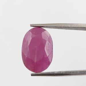 Könnte beinhalten: Ein facettierter, ovaler Rubin-Edelstein, gehalten von einer Pinzette. Der Rubin hat eine tiefe, gesättigte, rötlich-rosa Farbe mit sichtbaren inneren Strukturen und Facetten. Der Hintergrund ist schlicht weiß.