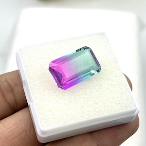 Può includere: Una gemma rettangolare sfaccettata caratterizzata da una vibrante sfumatura di colore che passa dal viola intenso a un'estremità al verde acqua brillante all'altra, esposta all'interno di un piccolo contenitore quadrato in plastica bianca con inserto in schiuma.