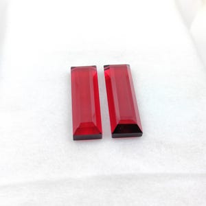 May include: Two rectangular, faceted, deep red gemstones. The gems have a polished, transparent appearance and are set against a white background. The shape is a long, slender rectangle with angled edges.