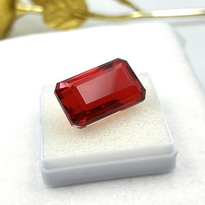 Può includere: Una gema rossa rettangolare sfaccettata con un aspetto chiaro e trasparente. La pietra presenta bordi netti e puliti e una superficie lucida, poggiata su un piccolo inserto in schiuma bianco di forma quadrata per l'esposizione.