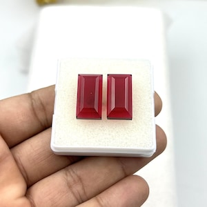 Puede incluir: Dos piedras preciosas rectangulares facetadas de color rojo intenso, dispuestas una al lado de la otra sobre una base de espuma blanca dentro de un pequeño recipiente de plástico cuadrado. Las piedras tienen una superficie pulida y brillante.